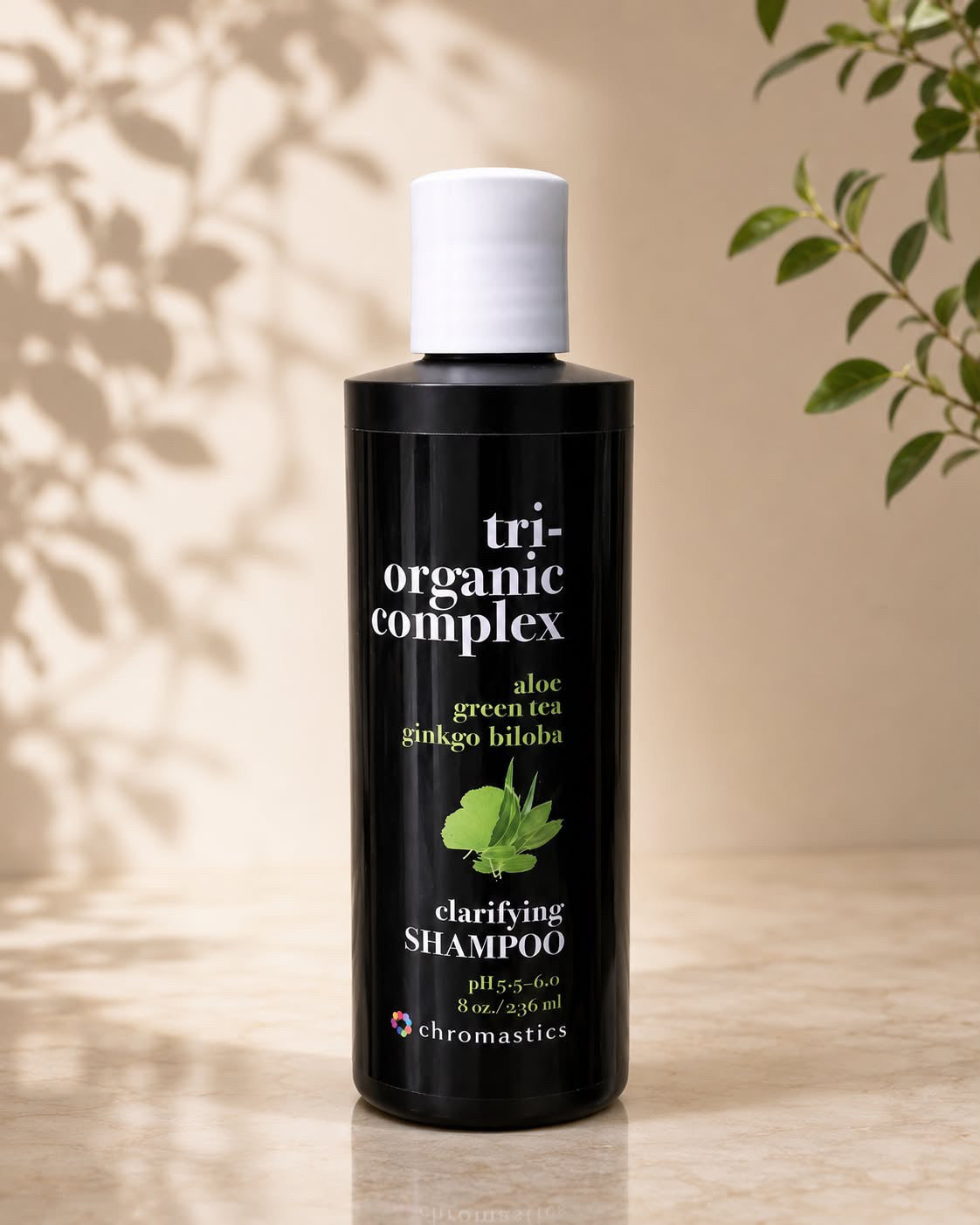 A bottle of Chromastatics Tri-Organic Complex Clarifying Shampoo, featuring a matte black bottle and white cap, set against a warm beige background with soft leaf shadows and green plant leaves.