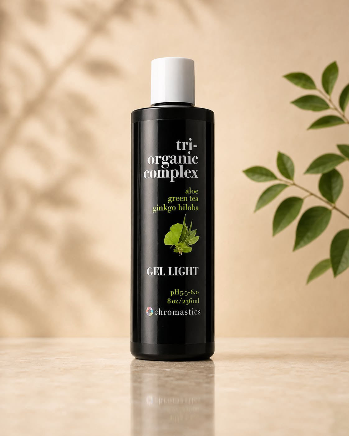 A bottle of Chromastics Tri-Organic Complex Gel Light skincare product, standing on a light beige surface with a warm beige background and soft leaf shadows, with a green leafy branch on the right.