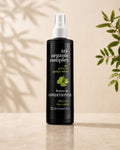 A black bottle of Chromastics Tri-Organic Complex Leave-In Conditioner with a white spray top, standing on a light beige surface against a warm beige background with soft leaf shadows and green leaves on the sides.