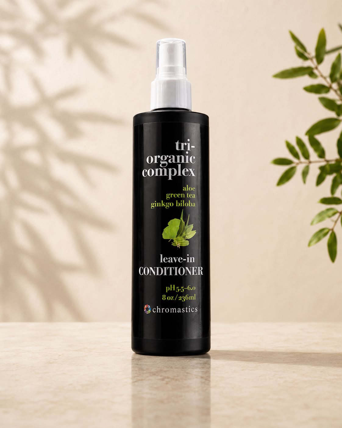 A black bottle of Chromastics Tri-Organic Complex Leave-In Conditioner with a white spray top, standing on a light beige surface against a warm beige background with soft leaf shadows and green leaves on the sides.