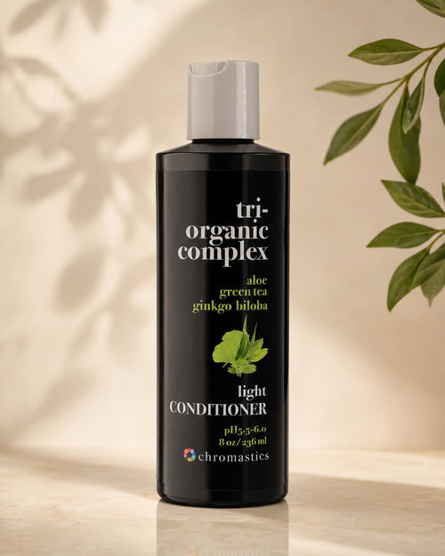 A bottle of Chromastics Tri-Organic Complex Aloe Green Tea Ginkgo Biloba Light Conditioner, standing on a light beige surface with soft leaf shadows and green leaves in the background.