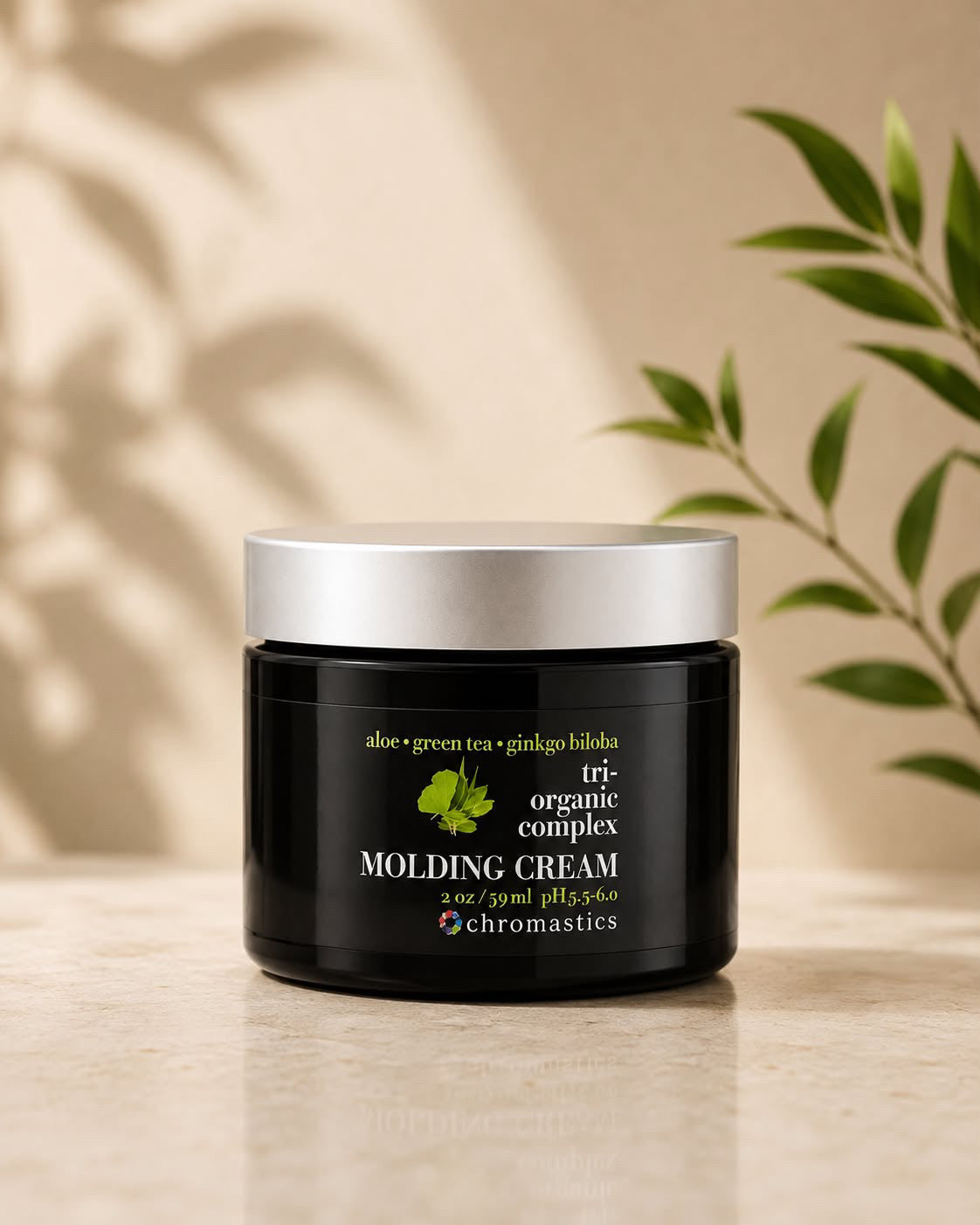A jar of Chromastics Tri-Organic Complex Molding Cream sits on a light stone surface with a beige background, featuring soft leaf shadows and green plant leaves framing the scene.