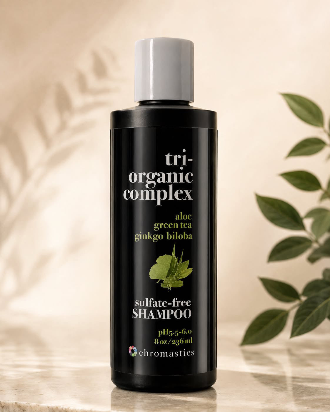 A bottle of Chromastics Tri-Organic Complex Sulfate-Free Shampoo, featuring a matte black bottle and a white cap, set against a warm beige background with soft leaf shadows and green plant leaves on the side.